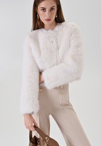 Weiße, kurze Jacke aus Kunstpelz mit weiten Ärmeln und strukturierter Stoffoberfläche. Kombiniert mit beigen, hoch taillierten Hosen mit goldfarbenen Knopfverzierungen.