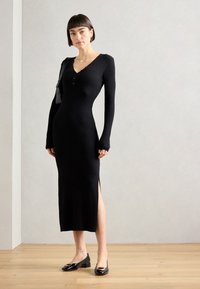 Abito in maglia nera a coste con maniche lunghe e scollo a V. Presenta uno spacco laterale e bottoni lungo il collo. Indossato con ballerine nere.