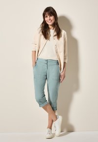 Světlý béžový button-up kabát přes bílý top, doplněný o mátově zelené zkrácené kalhoty a bílé tenisky. Hladká tkanina a ležérní střih.