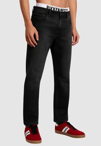 Jeans in denim nero con vestibilità dritta, dotati di cinque tasche. Abbinati a scarpe da ginnastica rosse e a una fascia bianca con scritte nere.