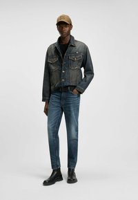 Jeansjacka med blekblå finish, klassisk krage, framfickor, i kombination med blå jeans och svarta gummistövlar, prydd med en beige keps.