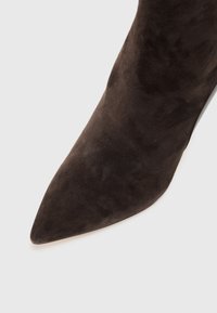 Botín de ante marrón con puntera afilada, de textura suave y silueta elegante, que presenta un tacón delgado y brillante, sin herrajes visibles.