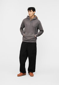 Νέος άνδρας που στέκεται με τα χέρια στις τσέπες, φορά γκρι φούτερ Timberland, μαύρο cargo παντελόνι και καφέ σουέτ παπούτσια σε λευκό φόντο.