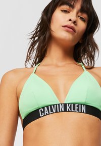 Šviesiai žalia trikampio formos maudymosi kostiumėlio viršutinė dalis su plonomis dirželiais ir juoda elastinga juosta, ant kurios yra balta "Calvin Klein" logotipas. Lygus paviršius.