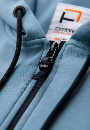Hellblauer Kapuzenpullover mit einem schwarzen Reißverschluss und Kordeln. Verfügt über ein Etikett mit der Aufschrift "OMBRE" in orangefarbenen Akzenten, das die glatte Textur des Stoffes hervorhebt.