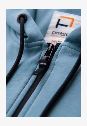 Haut à capuche bleu clair avec une fermeture éclair noire et des cordons de serrage. Présente une étiquette portant la mention "OMBRE" avec des accents orange, mettant en valeur la texture lisse du tissu.