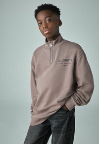 Felpa a zip corta color marrone chiaro con texture a coste e un piccolo logo, abbinata a jeans grigi scuri. Il modello si trova davanti a uno sfondo chiaro.