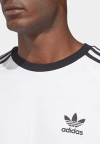 Biela bavlnená tričko s čiernym rebrovaným okrúhlym výstrihom, s tromi čiernymi pruhmi na pleciach a vyšitým čiernym logom Adidas.
