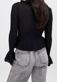 Sort tekstureret langærmet top med en flared ærmeudformning og rynket halsudskæring, parret med lys grå højtaljede denimjeans.