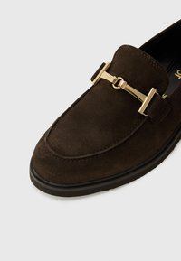 Braune Wildleder-Loafer mit einem goldfarbenen Kettenakzent, runder Zehenform und einer strukturierten Außensohle. Schlichtes Design mit minimalen Nähten.