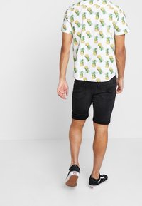 Camisa blanca de manga corta con estampado de piñas amarillas, combinada con pantalones cortos de mezclilla negros y zapatillas negras con suelas blancas, vista desde atrás.