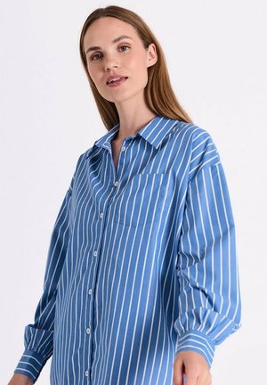 Vrouw die een loszittend blauw overhemd met witte verticale strepen en een borstzak draagt, staand voor een witte achtergrond.