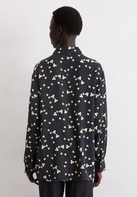 Zwarte shirt met wit bloemenpatroon, lange mouwen en een kraag. Glad materiaal met een losse pasvorm en knoopmanchetten.