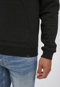 Svart hettegenser med kengurulomme og ribbestrikkede mansjetter. Bæres med lyseblå denimjeans. Lappen nederst leser "CASUAL ADVISOR".