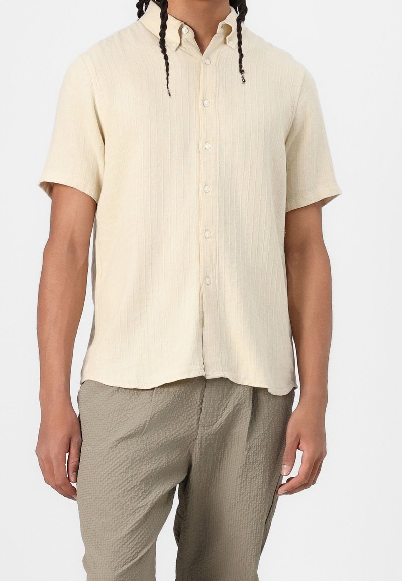 Kortærmet, lysebeige skjorte med knapper og ribbet struktur, med krave, fem knapper og en afslappet pasform. Bruges sammen med teksturerede grå bukser.