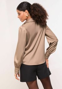 Long-sleeve, taupe satin blouse with a classic collar, puffed shoulders, and button accents on cuffs, paired with black shorts.