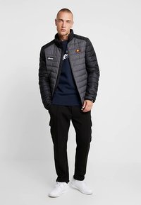 Casaco preto acolchoado com gola alta, com emblemas de logótipo, usado sobre uma sweatshirt azul-marinho e calças cargueiras pretas, combinado com ténis brancos.