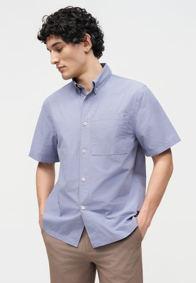 Άνδρας με σγουρά μαλλιά, που φορά ένα ανοιχτό μπλε κοντομάνικο πουκάμισο με κουμπιά και μπεζ παντελόνι, με τα χέρια στις τσέπες, κοιτάζει προς τα κάτω αριστερά.