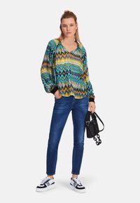 Multicolored long-sleeve blouse with geometric patterns, paired with blue skinny jeans and black and white sneakers, holding a black bag.
