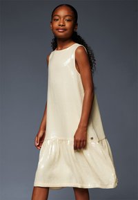 Robe sans manches couleur crème, ornée de paillettes, avec un col rond et un ourlet à volants en étages. Présente un petit accent logo sur le côté.