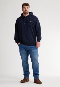 Felpa blu navy con cappuccio e lacci, con un piccolo logo. Abbinata a jeans azzurri e scarpe marroni. Design semplice e casual.
