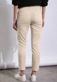 Pantalons cargo beiges à coupe slim avec des poches plaquées sur les côtés, associés à des baskets blanches. Tissu texturé, design longueur cheville.
