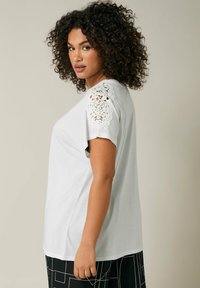 Top bianco a maniche corte con dettagli in pizzo floreale sulle spalle. Tessuto morbido, vestibilità rilassata, con scollo rotondo e orlo leggermente curvo.