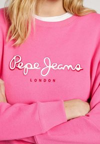 Žena v jasně růžovém svetru Pepe Jeans London se zkříženýma rukama, na kterém je logo v bílých a červených písmenech.