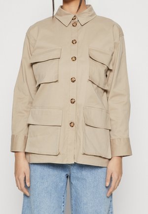 Veste utilitaire beige à boutons avec quatre grandes poches à rabat, portée avec un jean bleu clair, montrant le haut du corps et les mains.