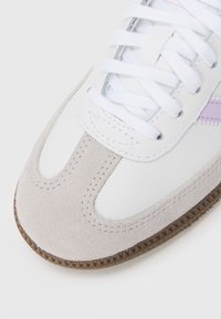 Baskets blanches avec une capuche en suede beige, des lacets blancs et une petite accentuation violette sur le côté, présentées sur un fond blanc uni.