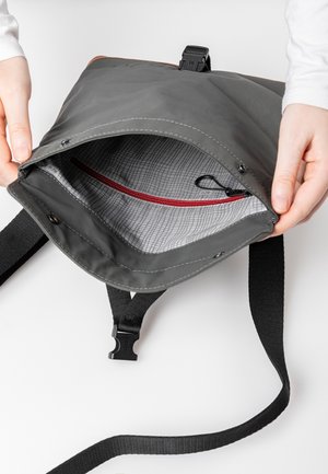 Bolsa impermeable gris con solapa frontal, correa negra y bolsillo interior con cremallera roja. Forrada con tela a cuadros, con cierre de clip.