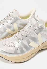 Ζευγάρι αθλητικών παπουτσιών σε ανοιχτό μπεζ και ασημί χρώμα με λεπτομέρειες από δίχτυ, μπροστινά κορδόνια και λογότυπα σε πλαϊνά και γλώσσες.