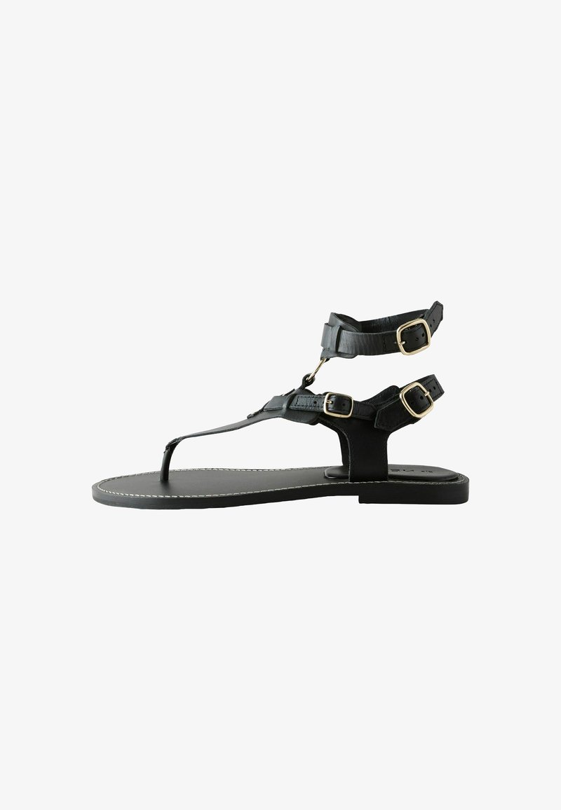 Sandales en cuir noir avec un design à bride en T, comportant deux brides ajustables à la cheville, des boucles dorées et une semelle plate avec des détails de couture blanche.