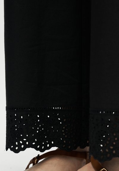 Μαύρο ύφασμα με λεπτομερή κεντήματα με κοψίματα στο τελείωμα πάνω από πόδια που φορούν καφέ δερμάτινα σανδάλια με χρυσά αγκράφες.