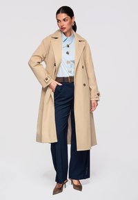 Femme portant un trench-coat beige sur une blouse bleu clair à gros boutons, un pantalon bleu marine à fines rayures, une ceinture marron et des talons marron.