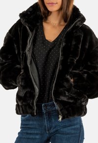Veste en fausse fourrure noire avec fermeture éclair, dotée d'un col haut et de poches latérales. Portée par-dessus un haut noir à pois et un jean en denim bleu.