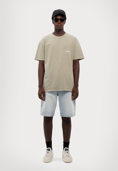 T-shirt de algodão verde-oliva com mangas curtas, combinada com calções de ganga azul-claro, meias pretas e ténis brancos. Boné preto e óculos de sol.