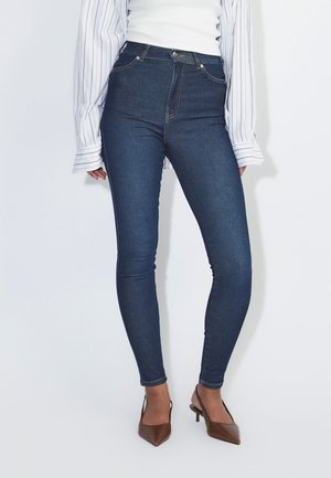 Mujer que lleva vaqueros pitillo azul oscuro, tacones marrones de punta afilada, una camiseta blanca y una camisa blanca de manga larga con rayas negras.