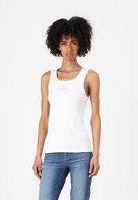 Weißes geripptes Tanktop mit breiten Trägern, auf der Front mit einem dezenten Logo versehen. Kombiniert mit blauen, figurbetonten Jeans. Schlichtes und figurbetontes Design.