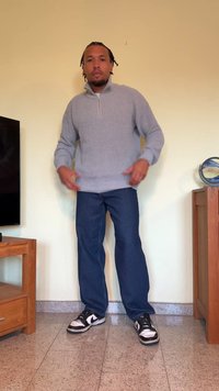 Sweater cinza de malha com meia abertura, calças de ganga azuis e ténis preto e branco. Em pé sobre um chão de azulejos, com móveis de madeira ao fundo.