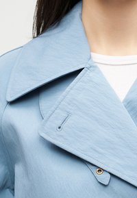 Κοντινό πλάνο ατόμου που φορά ανοιχτό μπλε υφασμάτινο μπουφάν με φαρδιά γιακά και χρυσή λεπτομέρεια με κόπιτσες πάνω από λευκό ζακάρ τοπ.