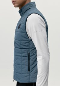 Gilet imbottito di colore blu ardesia con colletto alto, dotato di una tasca laterale con zip e un patch con logo. Indossato sopra una camicia a maniche lunghe bianca.