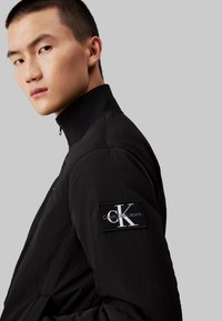 Melna viegla jaka ar augstu apkakli un teksturētu apdari. Izteiksmīgs pleca uzlīme atspoguļo Calvin Klein Jeans logo.