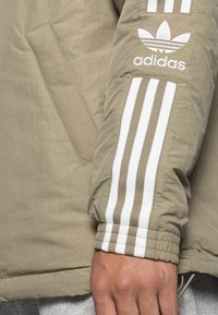 Beige jacka med en slät textur, som har en vit Adidas-logotyp och tre vita ränder på ärmarna. Inkluderar en sidficka.