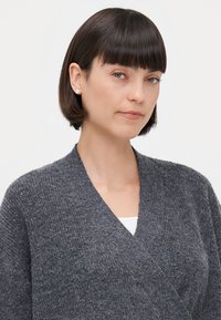 Grau gestrickte Strickjacke mit V-Ausschnitt, ausgestattet mit einem strukturierten Stoff und einem weichen, gemütlichen Aussehen. Getragen über einem weißen Hemd.