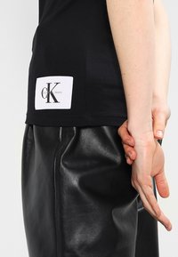 T-shirt noir avec un patch rectangulaire blanc affichant le logo "cK Calvin Klein Jeans". Le modèle porte un pantalon en cuir noir.