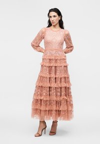 Γυναίκα με ροζ δαντελένιο φόρεμα με μακριά μανίκια και επίπεδα βολάν που φτάνει ως το πάτωμα, στέκεται με τα χέρια στη μέση και φοράει πέδιλα με τακούνια.