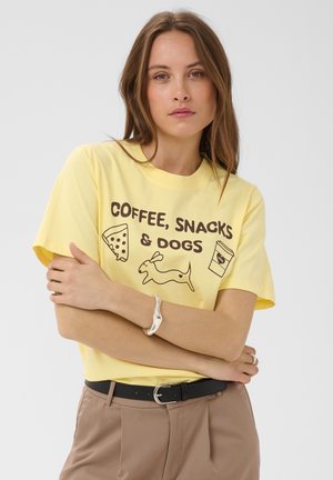 Femme aux longs cheveux bruns portant un t-shirt jaune avec le texte "COFFEE, SNACKS & DOGS" et des dessins de pizza, d'un chien et d'une tasse de café.