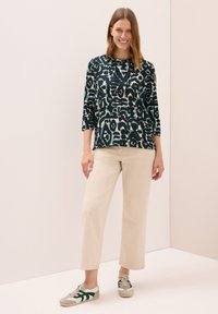 Blusa verde y negra con estampado, de mangas tres cuartos, combinada con pantalones rectos color beige claro y zapatillas con acentos verdes.