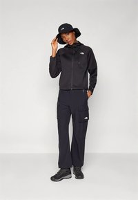 Γυναίκα σε μαύρο εξωτερικό ένδυμα με καπέλο, μπουφάν με φερμουάρ, cargo παντελόνι και μποτάκια πεζοπορίας, που στέκεται μπροστά από έναν ουδέτερο φόντο.